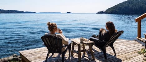 To personer sidder ved et træbord på en terrasse med udsigt over vand og fjerne kystlinjer.
