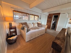 Ein gemütliches Zimmer mit einem weißen Sofa, einer Holzbank, einer Lampe und einem Fenster mit Jalousien.