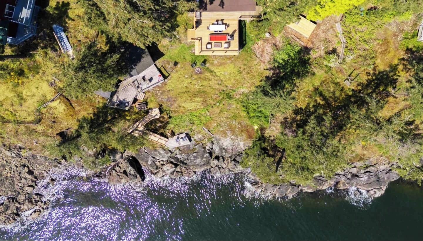 An aerial view of a coastal property with a wooden deck, a small building with a red roof, and a rocky shoreline.