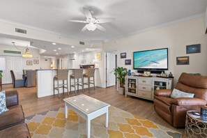 A modern living room with a television, seating, a kitchen area, and a ceiling fan. - Luxury Beachfront 2BR Sleeps 8 Pool Amazing Views (Panama City Beach)