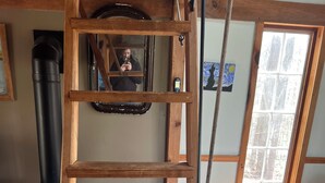 A wooden ladder, a sofa, a patterned rug, a fireplace, and a window with a view of trees. - Tiny Timber frame 1-bedroom cabin in Candor with AC/Heat, Outdoor fire and tub. (Candor)