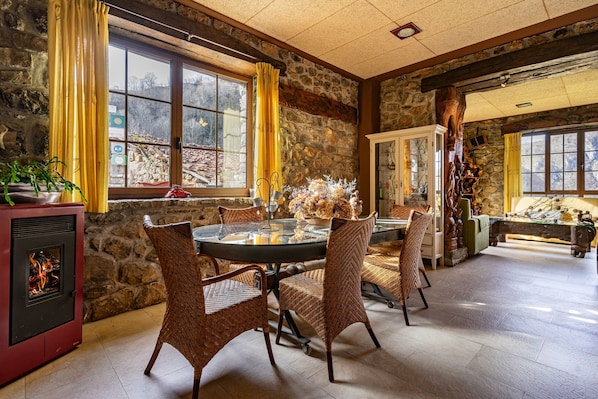 A stone-walled room with a fireplace, wicker chairs, a glass table, and a view through large windows.