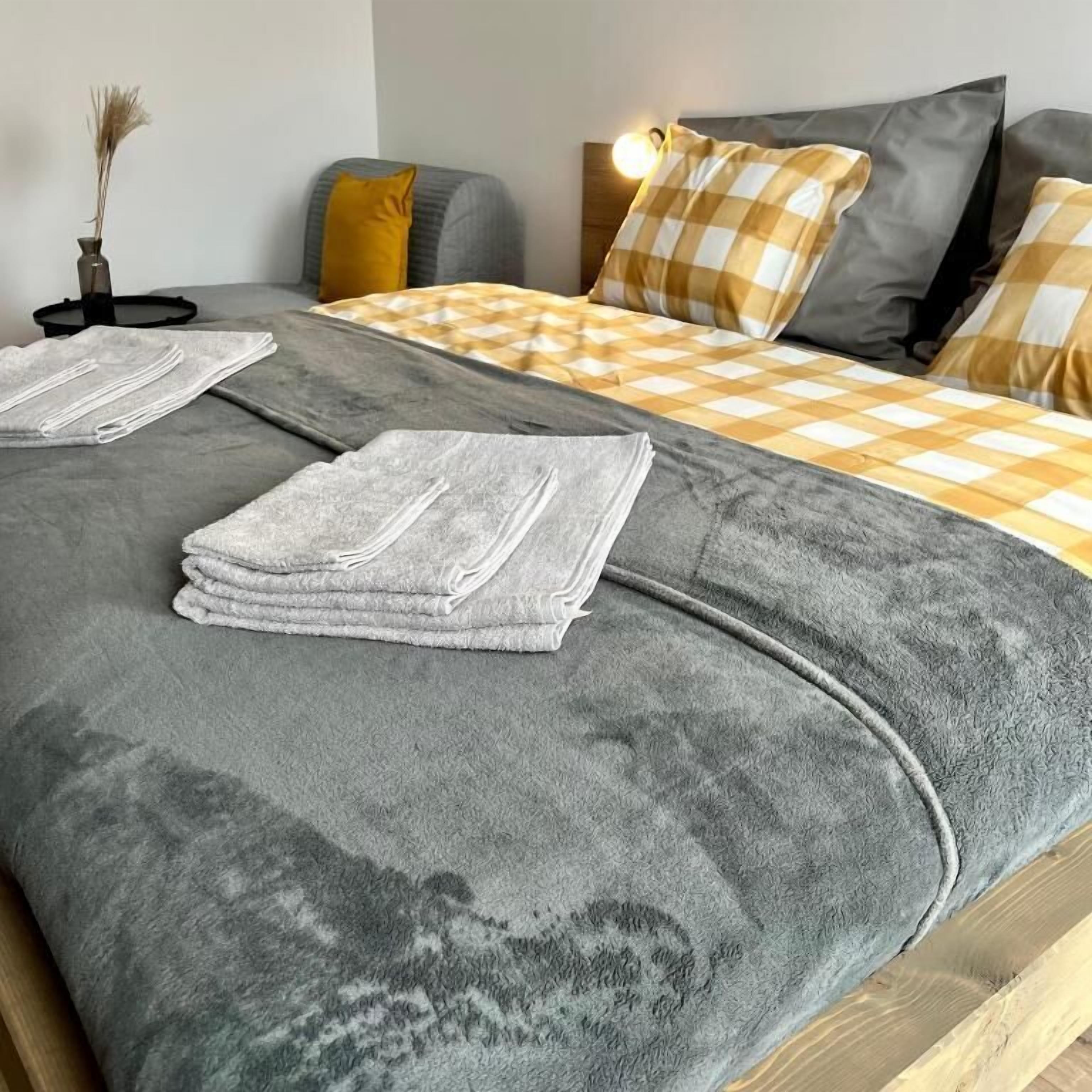 Un letto rifatto con un copriletto grigio e due asciugamani piegati, un comodino con una lampada e un vaso a parete con piante essiccate.