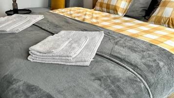 Un letto rifatto con un copriletto grigio e due asciugamani piegati, un comodino con una lampada e un vaso a parete con piante essiccate.