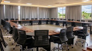 Un salón de conferencias con una larga mesa, sillas negras y ventanas amplias con cortinas.