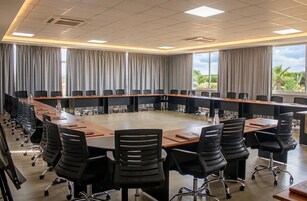Un salón de conferencias con una larga mesa, sillas negras y ventanas amplias con cortinas.