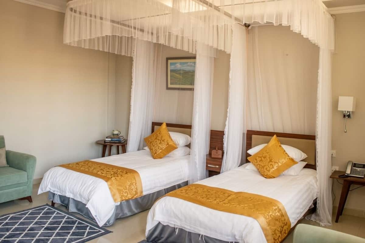A hotel room with two beds, a green armchair, a small table, and a painting on the wall.