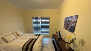 A bedroom with a bed, a dresser, a window with blinds, and a painting on the wall. - Room-a Peaceful and Scenic Area Designed (Cape Town)