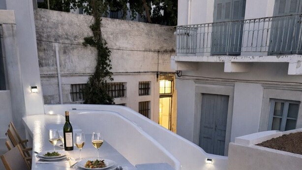 A modern outdoor dining area with a white table and chairs, a bottle of wine, and a glass of white wine. - Naxos Damarionas Retreat | Newly Renovated Cycladic House with Views (Damarionas)