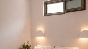A bedroom with a bed, bedside tables, a nightstand, a potted plant, a wall-mounted lamp, and a window. - Naxos Damarionas Retreat | Newly Renovated Cycladic House with Views (Damarionas)
