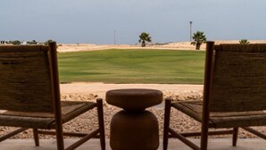 A view from a room with a wooden chair and a small table, overlooking a green field and a sandy area with palm trees. - Good Days Habitat Soma Bay (Soma Bay)