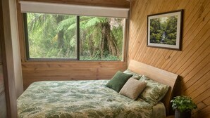 Habitació amb llit, finestra que dona a vegetació, quadre al mur i taula petita amb una planta.