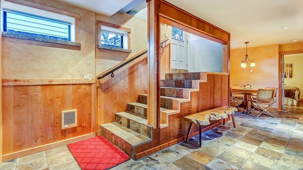 Una escalera de madera con un tapete rojo al final, un comedor con mesa y sillas, y vistas al exterior a través de una ventana.