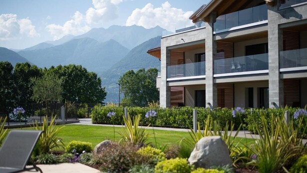 Un bâtiment moderne à deux étages, doté de balcons, entouré d’un jardin bien entretenu et avec des montagnes en toile de fond.