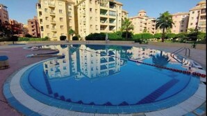 En pool med cirkelformad design, omgiven av en poolterrass med ett säkerhetsräcke. I bakgrunden syns fler våningar höga bostadshus.