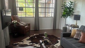 Ein Wohnzimmer mit Sofa, Sessel, Couchtisch, Deckenventilator und einem großen Fenster mit Vorhängen.