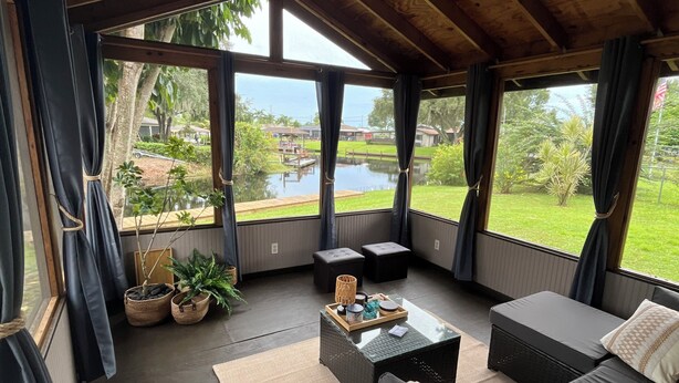Ein geräumiges Zimmer mit Deckenventilator, großen Fenstern, einer Couch, einem Couchtisch und Blick auf einen See und Grünflächen.