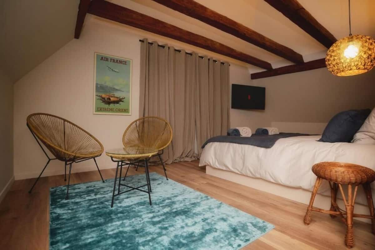 A bedroom with a bed, two wicker chairs, a blue rug, wooden beams, a poster, and a TV.