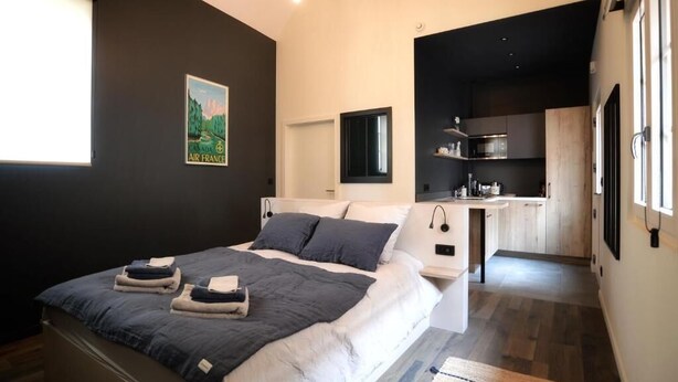 A modern bedroom with a bed, bedside tables, a small desk, and a view of a kitchen.