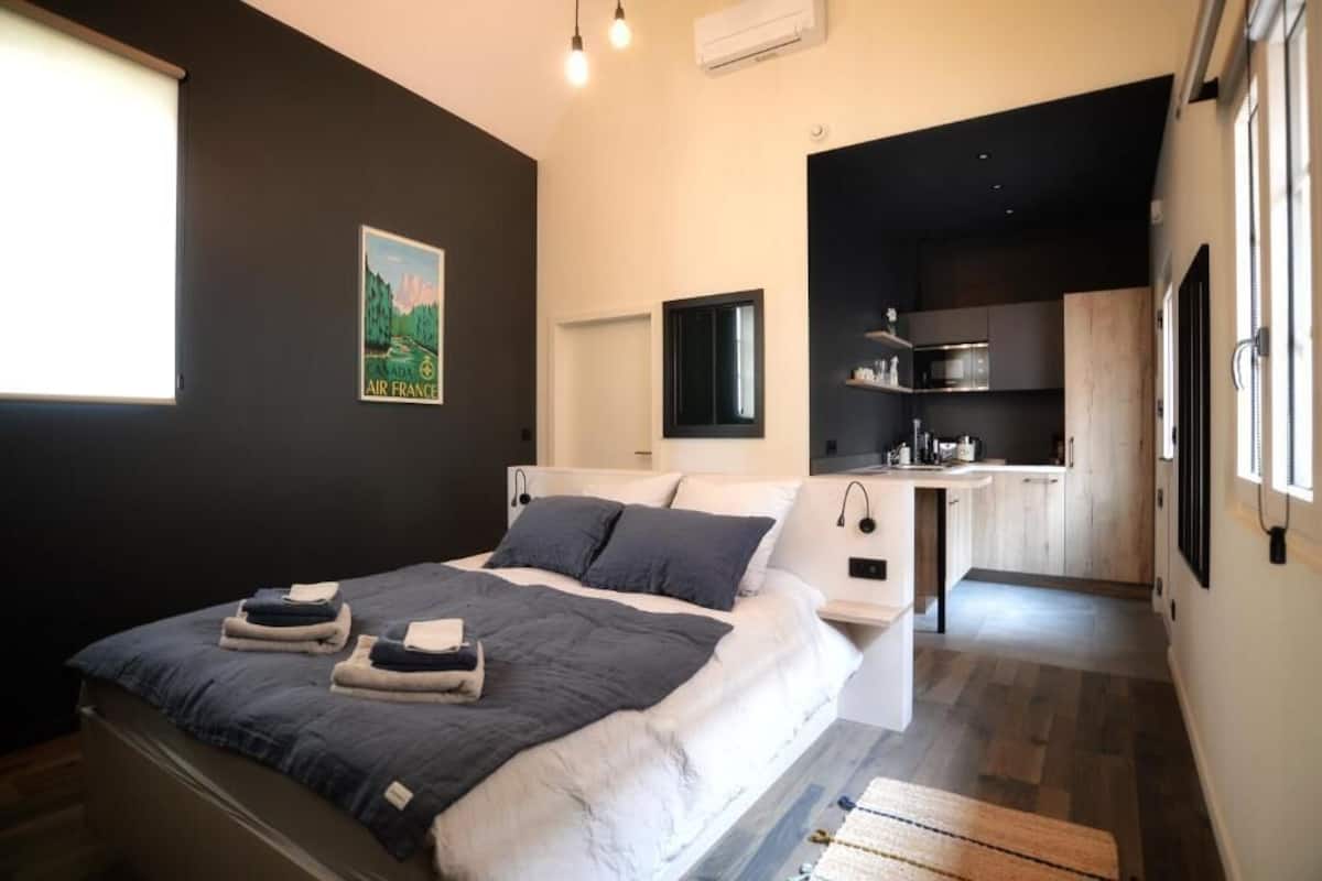 A modern bedroom with a bed, bedside tables, a small desk, and a view of a kitchen.