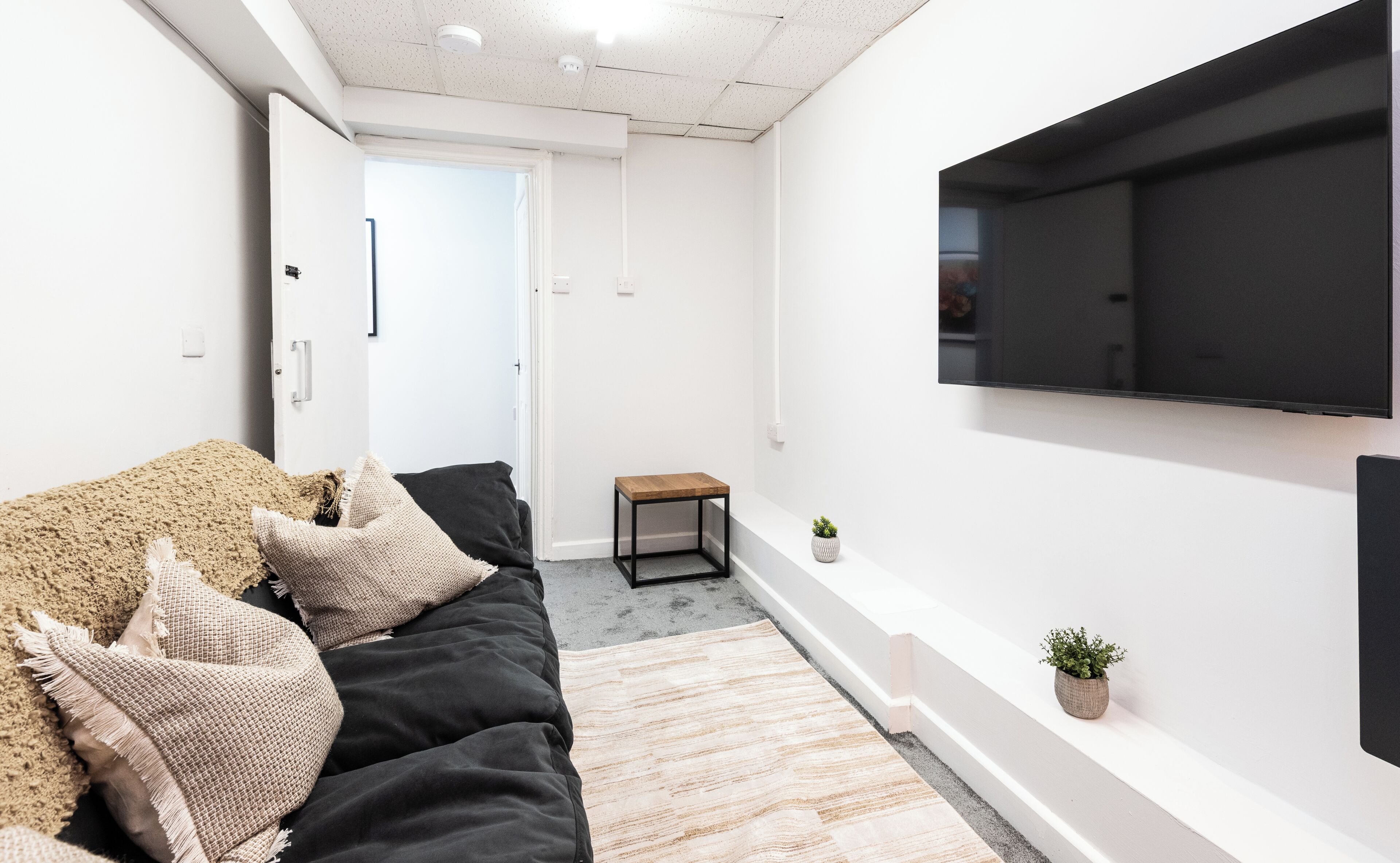 A modern living room with a black sofa, a small wooden side table, a mounted TV, and a potted plant.