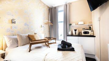 A hotel room with a bed, a wooden bedside table, a TV mounted on the wall, and a kitchenette area.