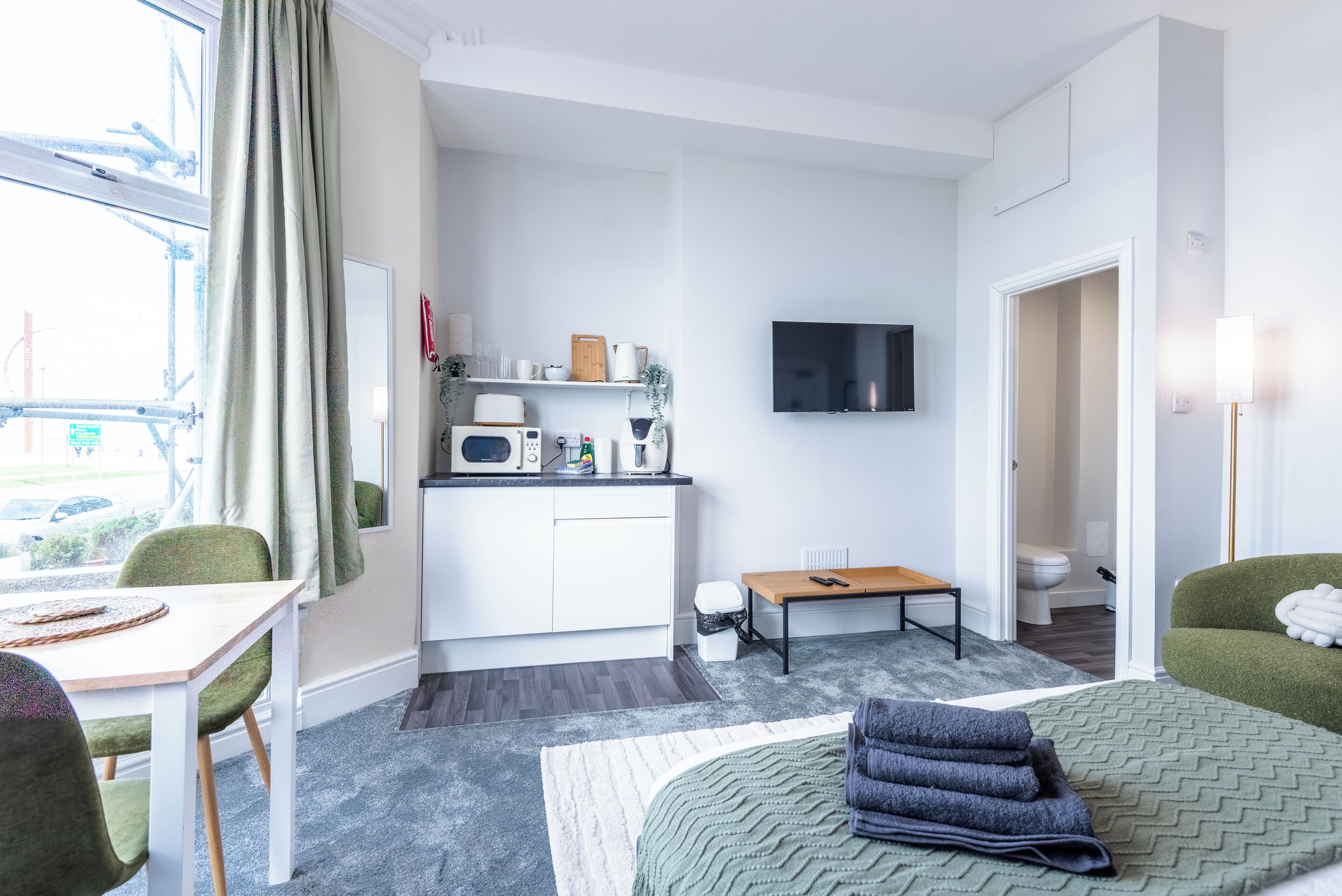 A compact hotel room with a kitchenette, a bed, a dining area, and a TV.
