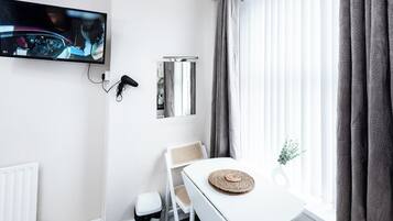 A room with a TV mounted on the wall, a white table with a woven placemat, a white chair, a radiator, and a window with curtains.