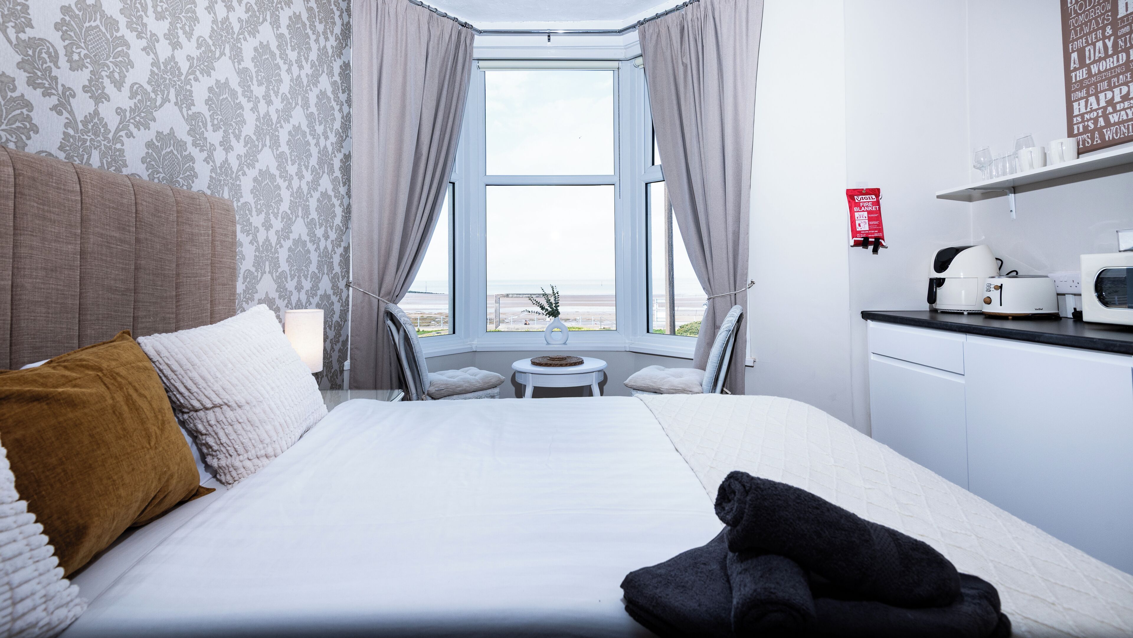 Habitación de hotel con cama, una mesita, una silla, vista a la playa, zona de cocina y una manta ignífuga en la pared.