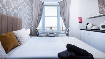 Habitación de hotel con cama, una mesita, una silla, vista a la playa, zona de cocina y una manta ignífuga en la pared.
