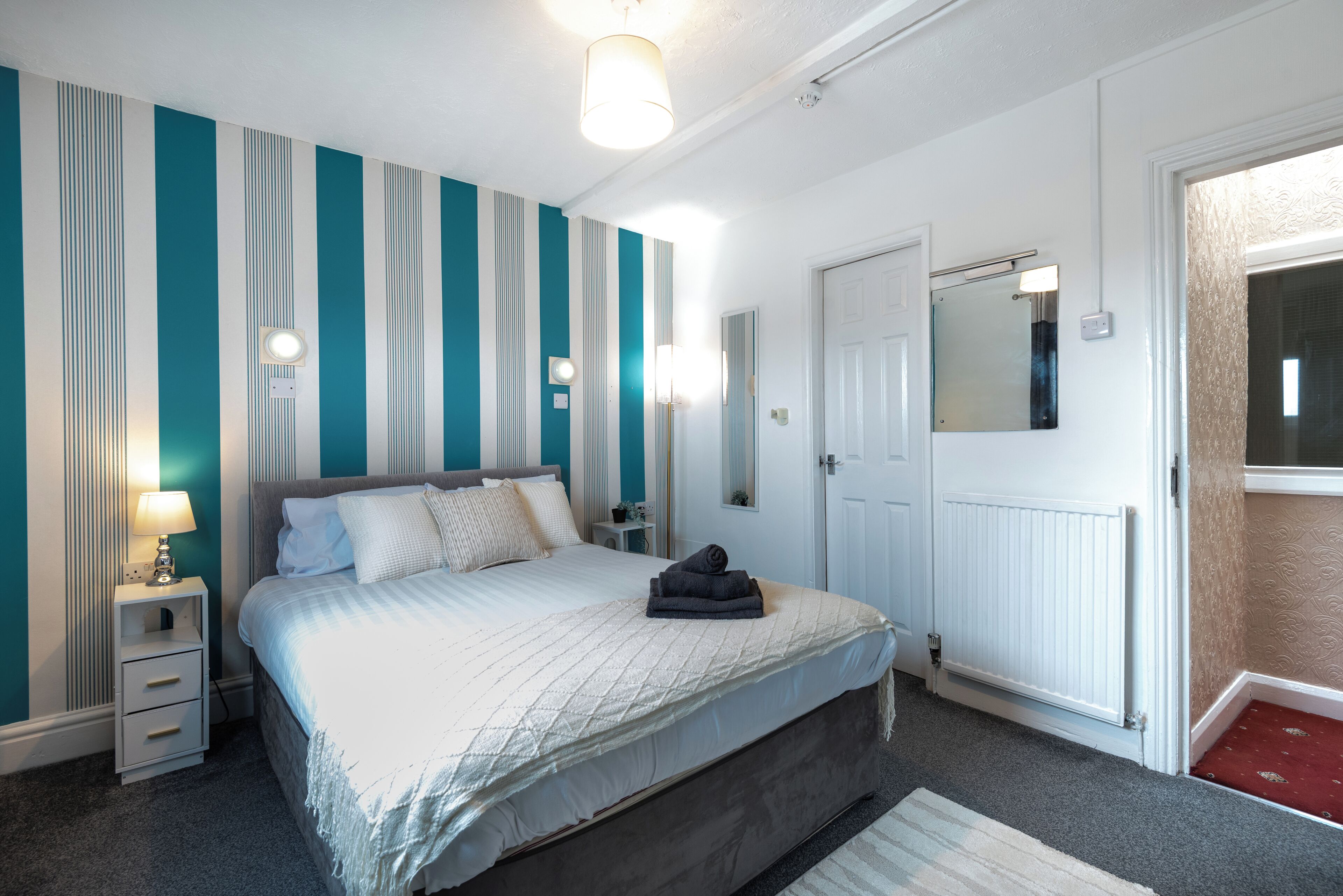 Un dormitorio con cama, mesita de noche, lámpara, espejo en la pared y una puerta con radiador.