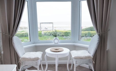 A corner room with two white wicker chairs, a small white table, and a view of the sea through large windows.