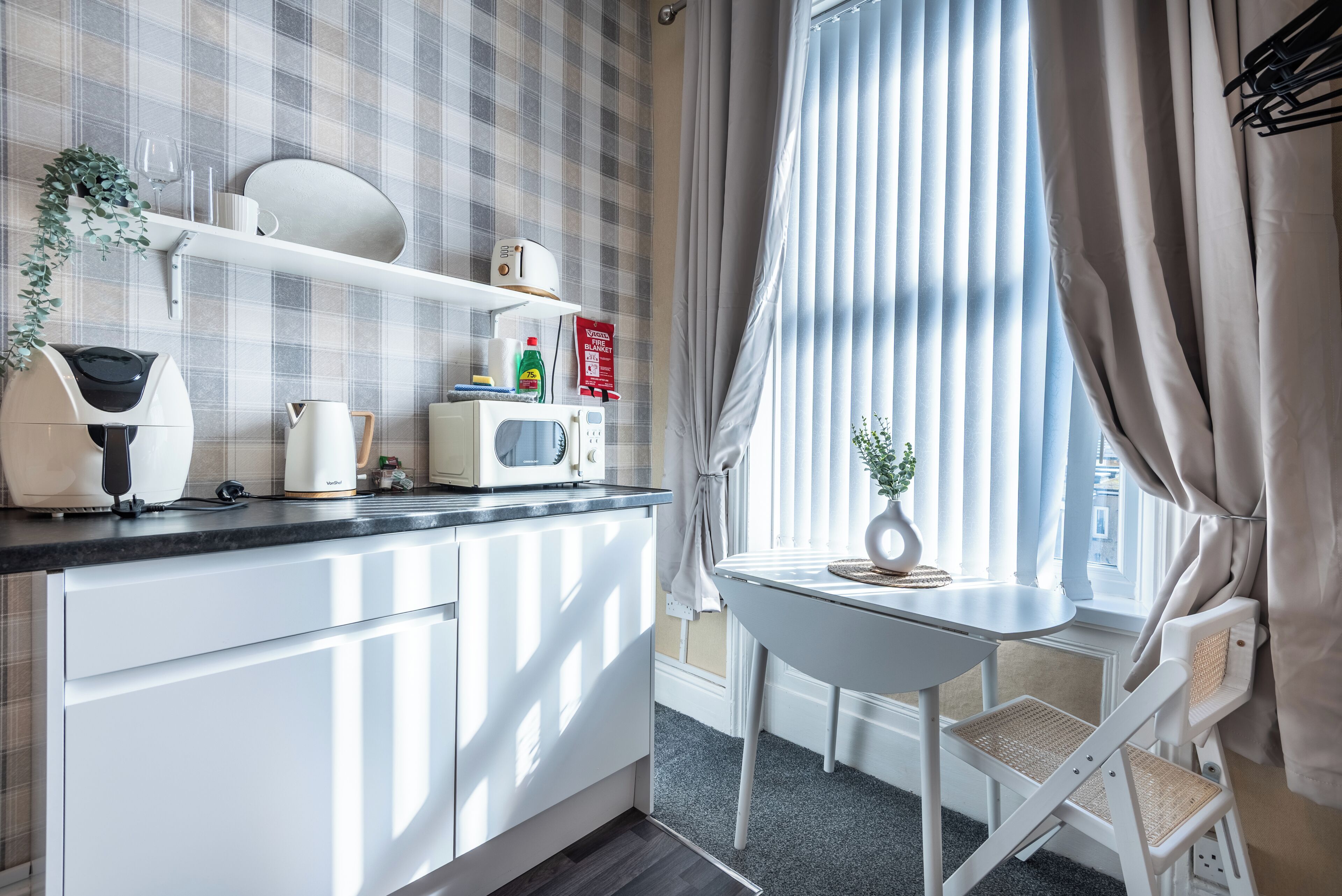 Een keuken met witte kastjes, een magnetron, een waterkoker en een klein tafeltje met een stoel.