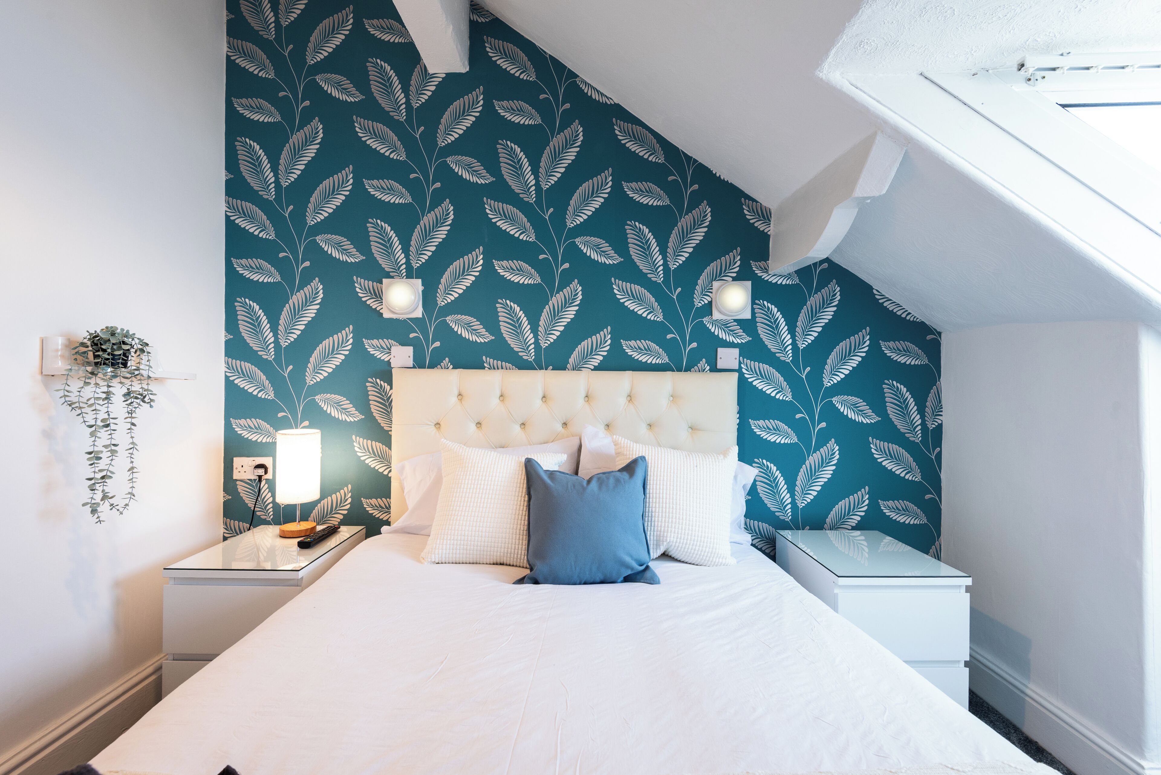 Un dormitorio con cabecera capitonada, cama con ropa de cama blanca y azul, dos mesitas de noche y un papel tapiz con estampado de hojas.