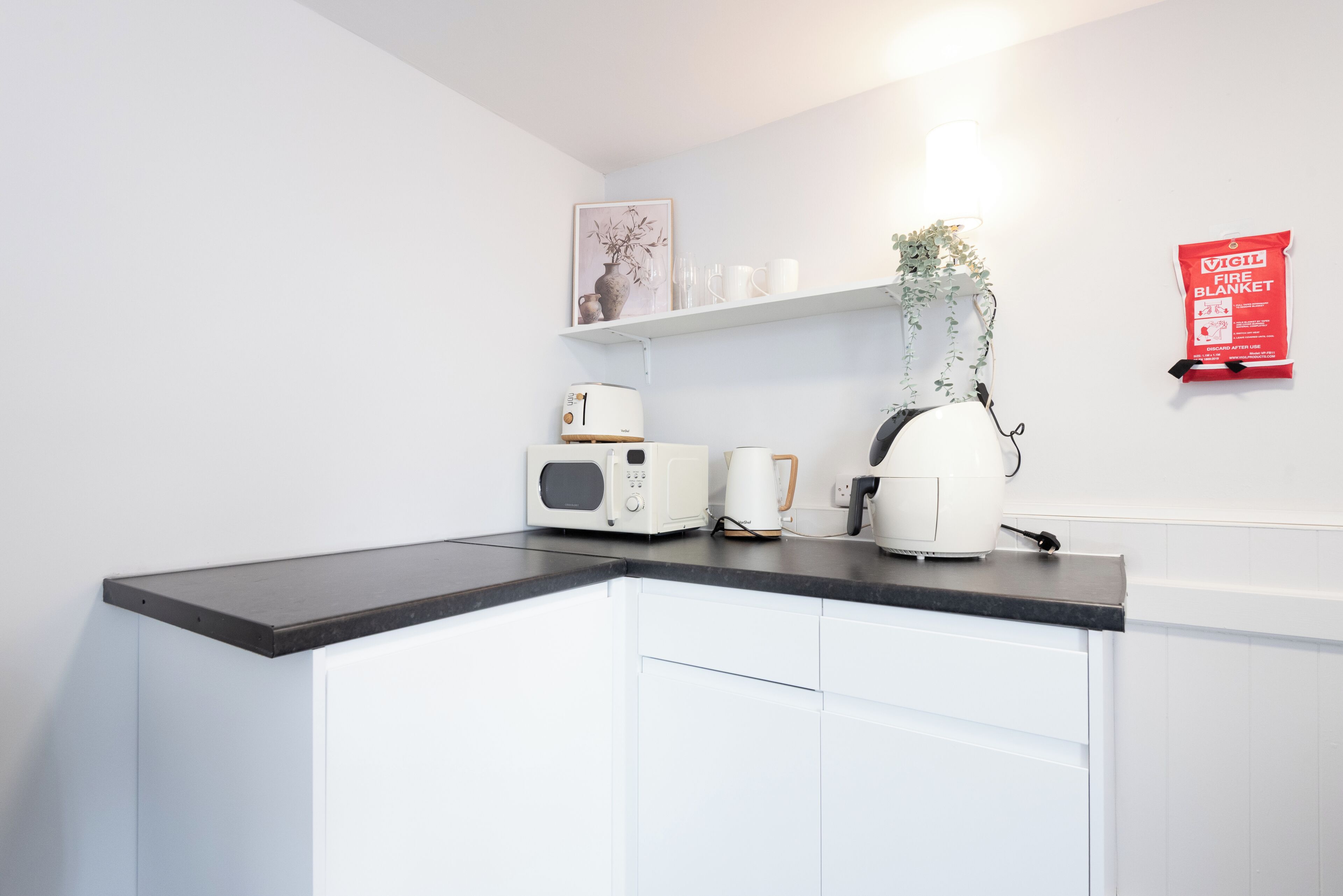 A kitchen with white cabinets, a black countertop, a toaster, a microwave, a kettle, a plant, a framed picture, and a fire blanket on the wall.