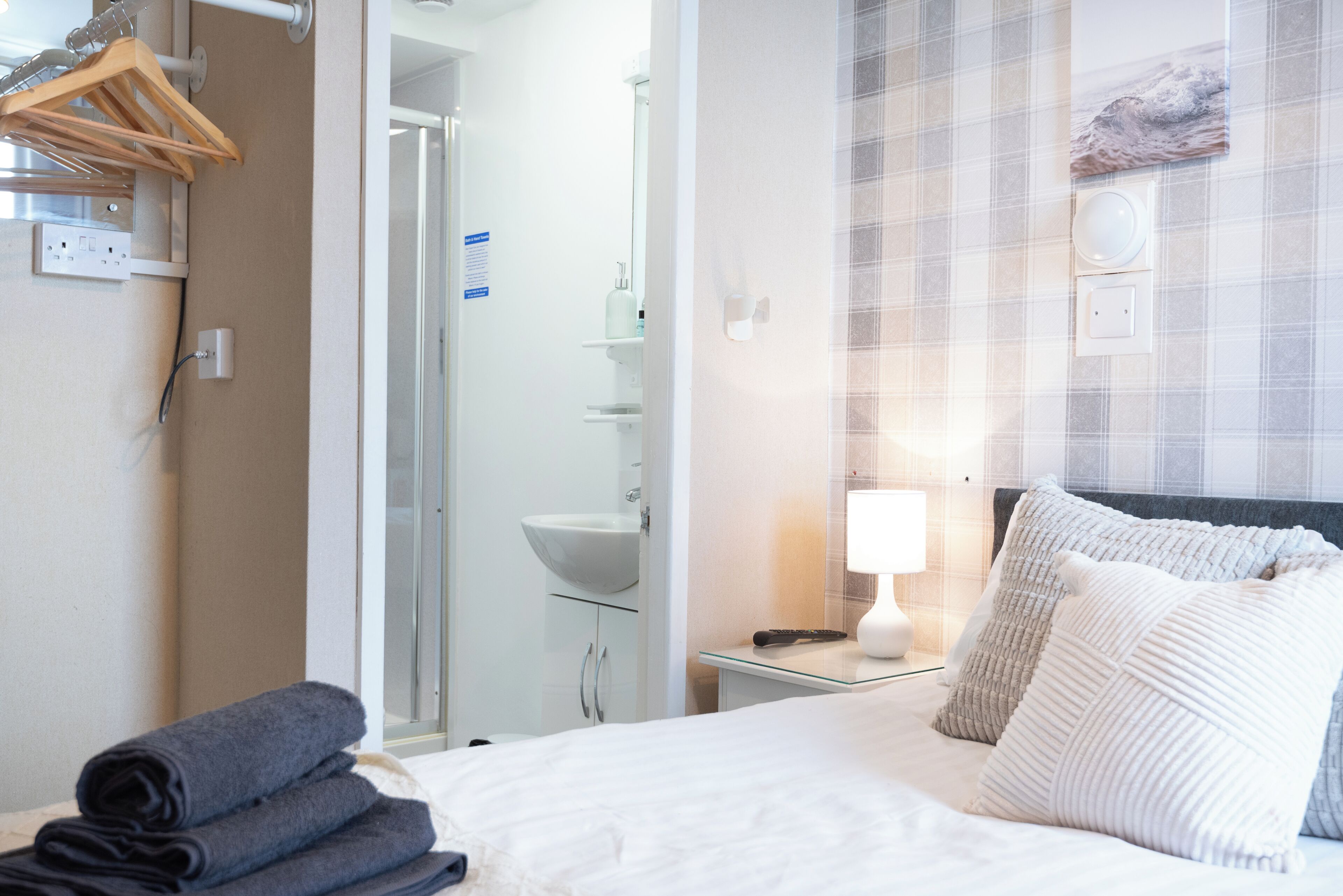 Habitación de hotel con una cama, una mesita de noche con lámpara, un interruptor de luz en la pared, un perchero para toallas y un baño visible a través de una puerta abierta.