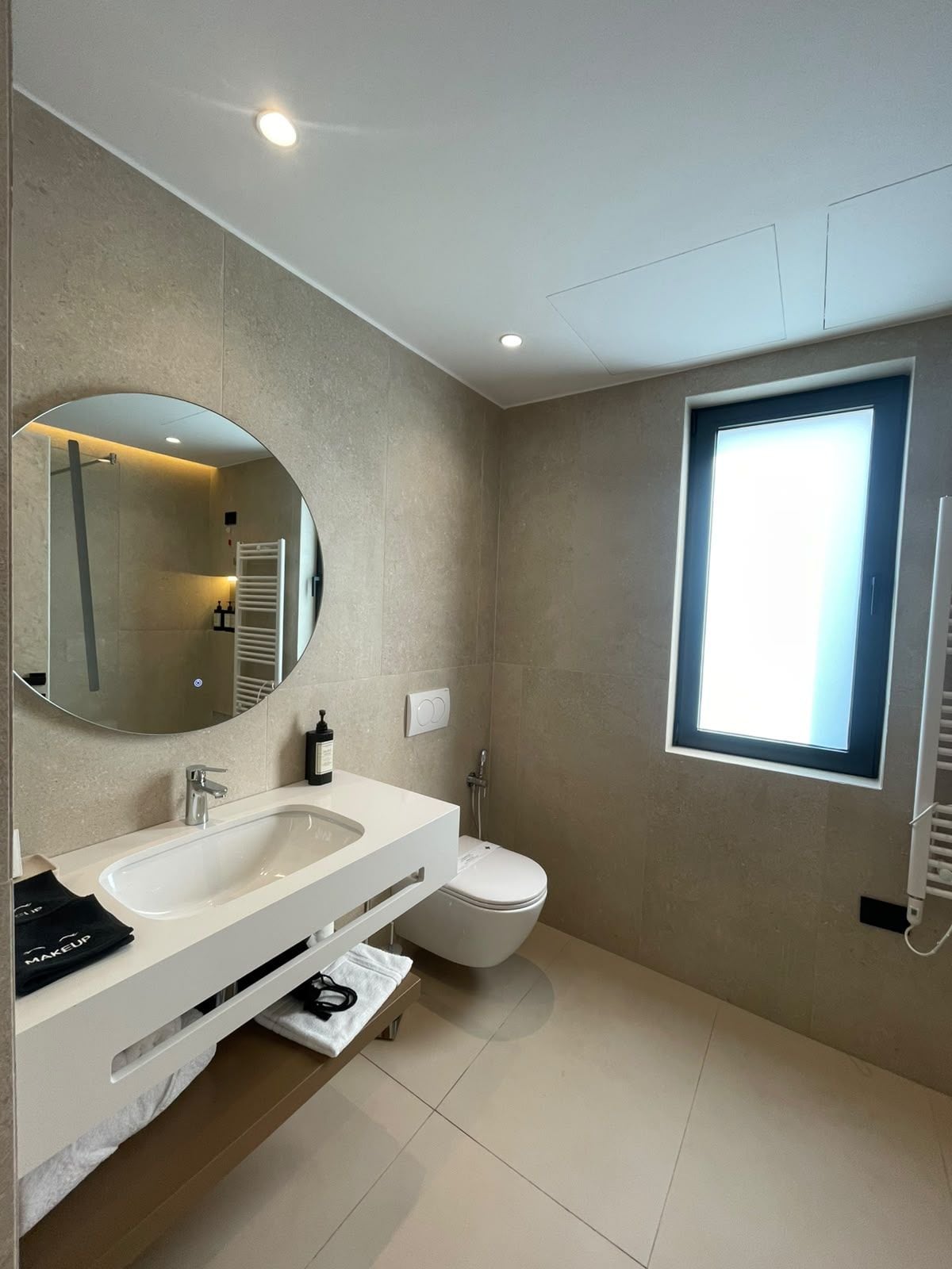 Un baño moderno con un espejo grande, un lavamanos, un inodoro y una ventana que deja entrar luz natural.