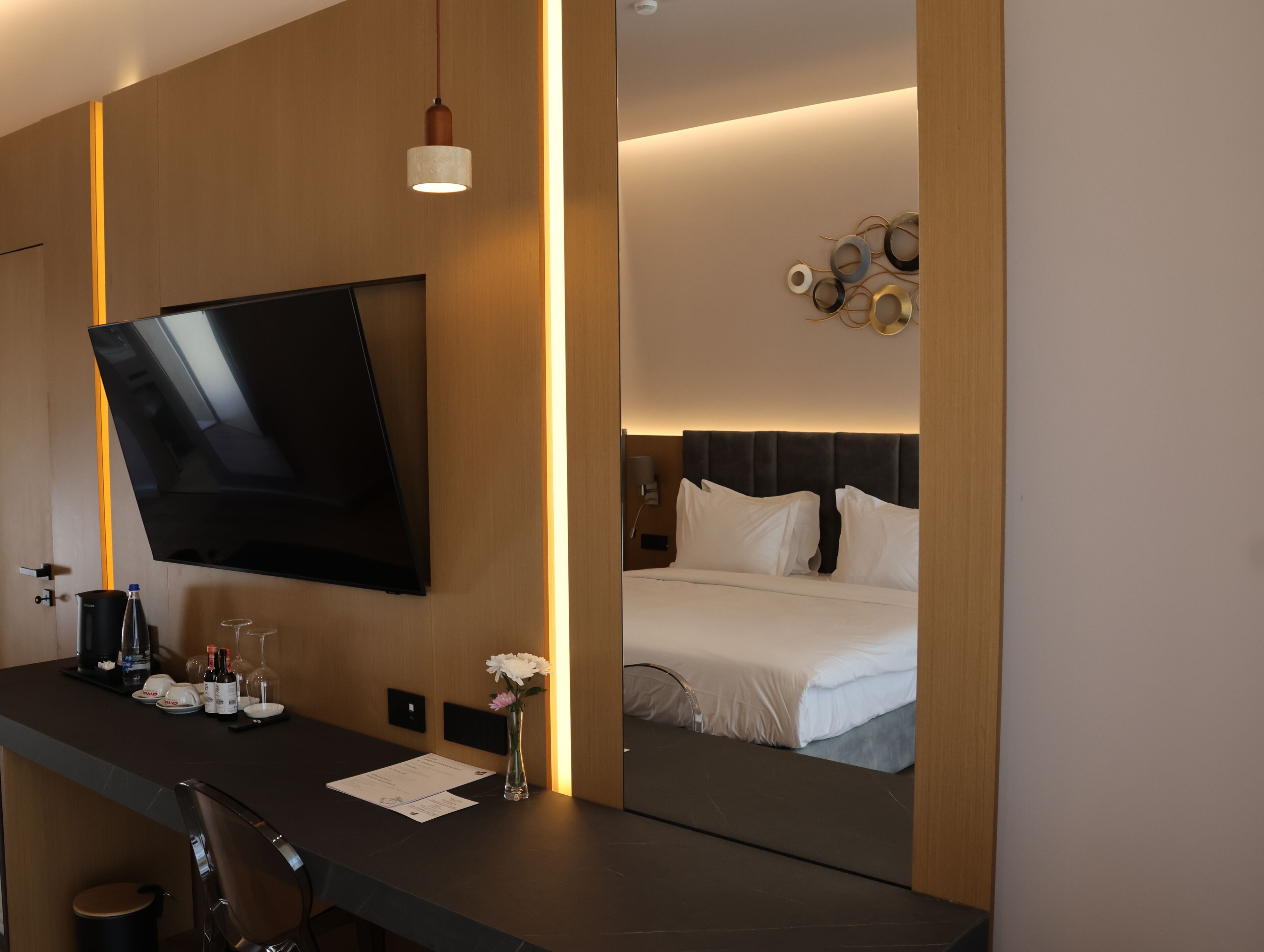 A hotel room with a flat-screen TV mounted on the wall, a bedside table with a vase of flowers, a bed with white linens, and a wall-mounted artwork.