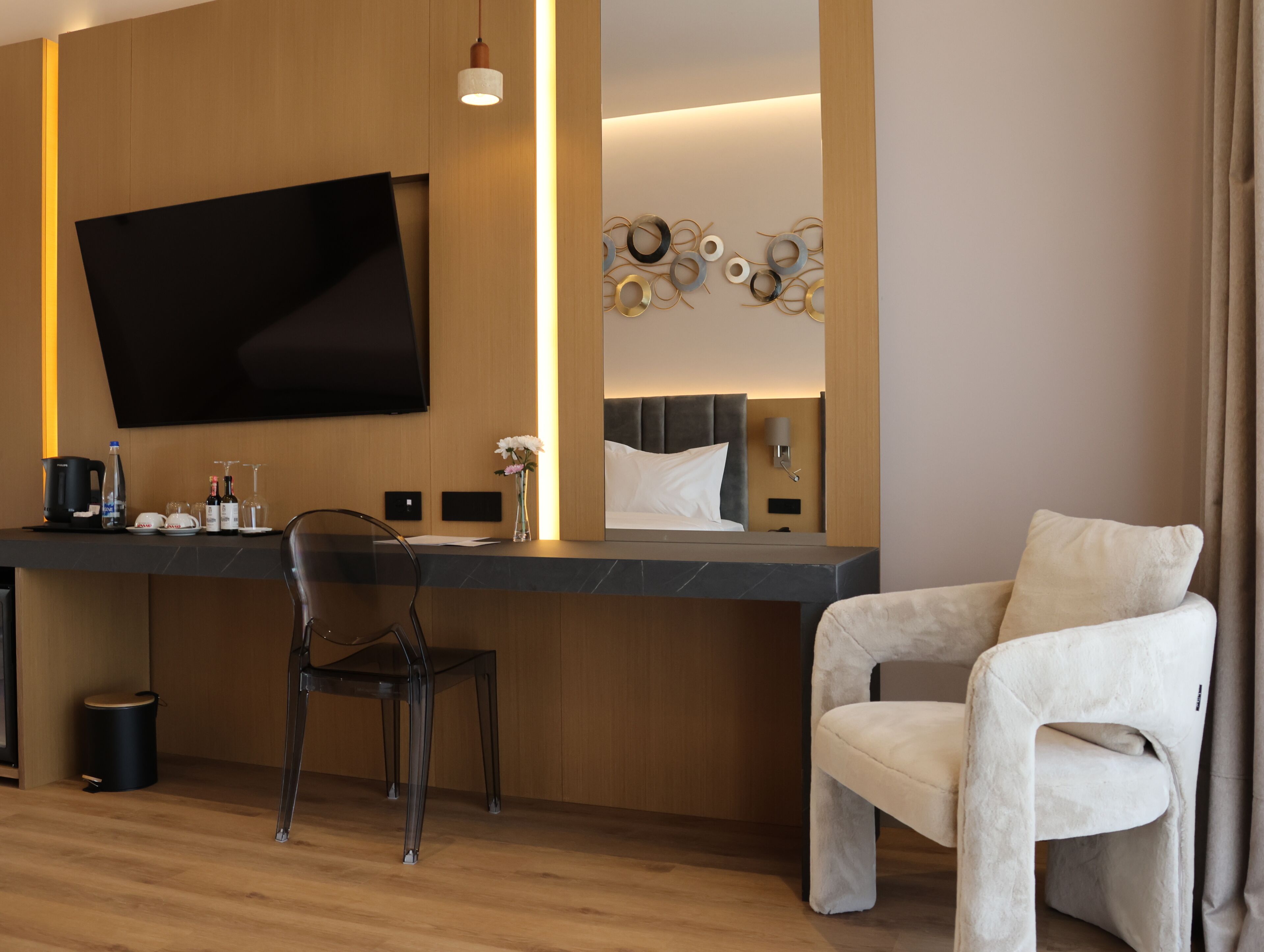 A modern hotel room with a flat-screen TV, a wooden accent wall, a decorative mirror, a comfortable armchair, and a dining area with a chair and table.