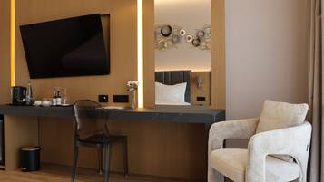 A modern hotel room with a flat-screen TV, a wooden accent wall, a decorative mirror, a comfortable armchair, and a dining area with a chair and table.