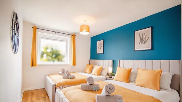 Hotellihuone, jossa on kaksi sänkyä, ikkuna verhoilla, sininen tehosteseinä ja taideteoksia.