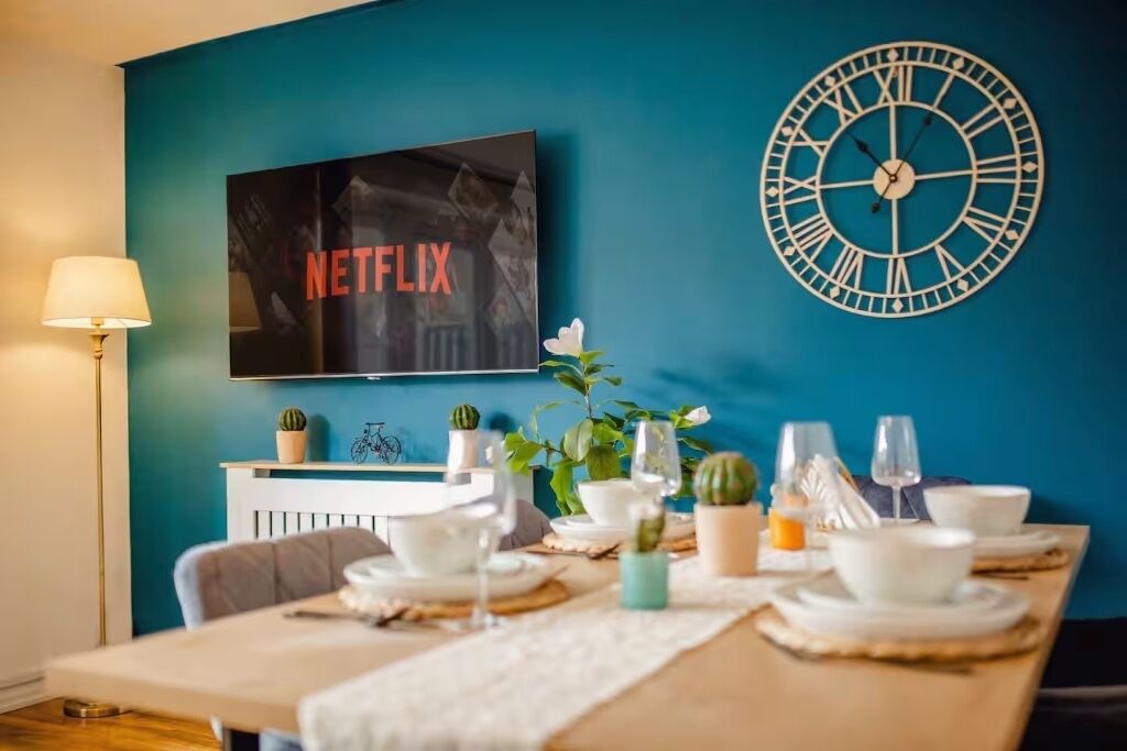Un'area pranzo con un tavolo apparecchiato per quattro persone, una sveglia e una TV che mostra 'NETFLIX' a parete.
