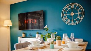 Ruokailutila, jossa on neljälle katettu ruokapöytä, seinäkello ja televisio, jonka näytöllä näkyy teksti ’NETFLIX’.