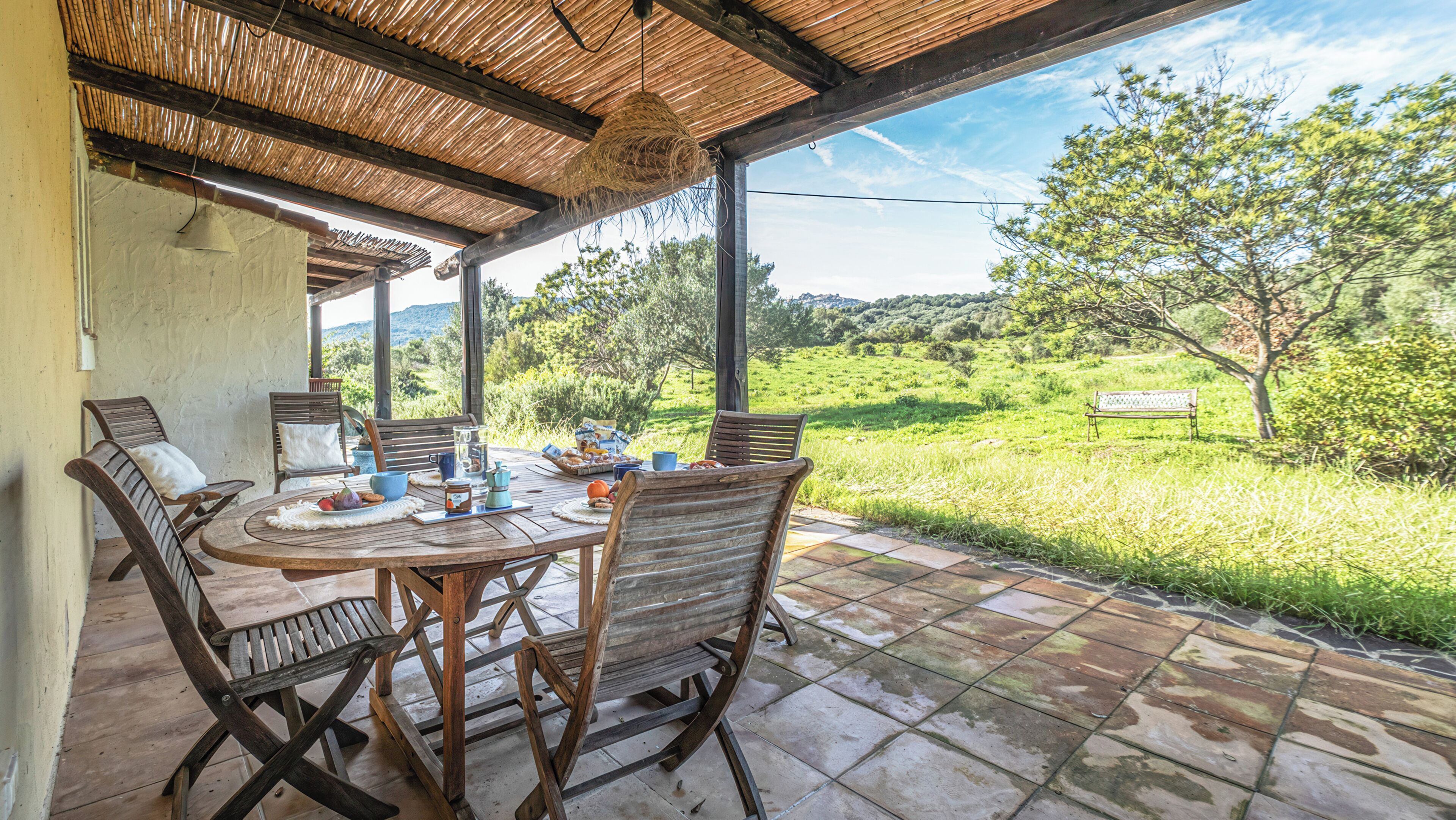 A covered patio with a table set for breakfast, surrounded by chairs, overlooking a grassy landscape with trees and a bench.