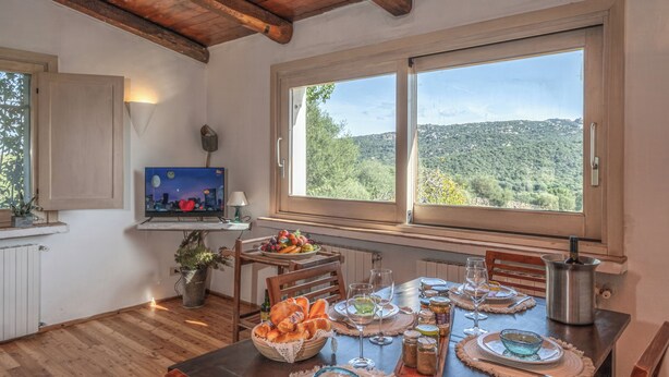 Un comedor con una mesa dispuesta para una comida, un televisor en un estante y vistas a un paisaje boscoso a través de amplias ventanas.