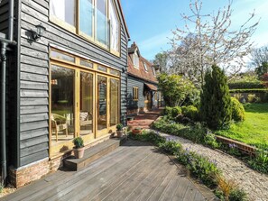 A house with a wooden exterior, large glass doors, a brick pathway, and a garden with various plants and a tree. - Six Apples (Braintree)