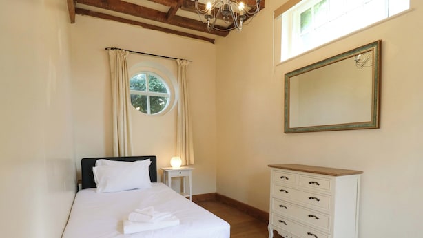 Ein Schlafzimmer mit Bett, Nachttisch, Kommode, einem Fenster mit Vorhängen und einer Kristallleuchter.