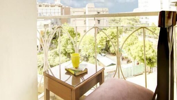 A balcony with a view of buildings and greenery, a table with a yellow cup, and a brown chair. - Marks Hotel (Noida)