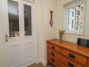 A room with a white door, a wooden dresser with drawers, a wall clock, and a window with blinds. - Kittiwake at No 20 (Seahouses)