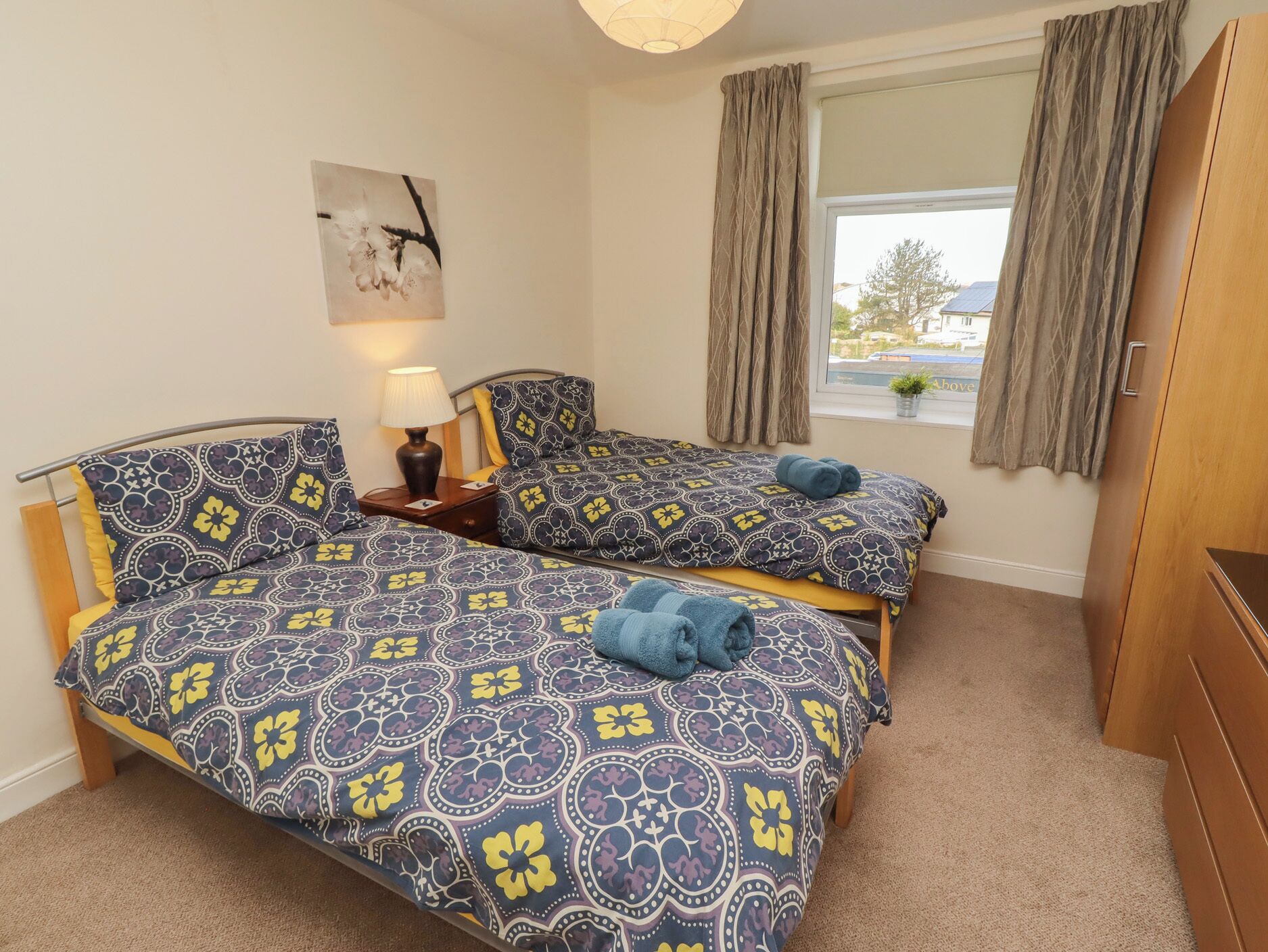 A bedroom with two beds, a bedside table with a lamp, a window with curtains, and a wall-mounted picture.